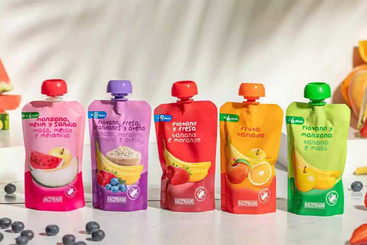 Cinco bolsitas de puré de fruta Hacendado de distintos sabores sobre una superficie blanca, con arándanos y fruta alrededor